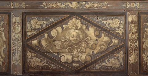 Panneau de bas-lambris à compartiment en losange inscrit dans un rectangle. Dans le losange, tête féminine, fleurs, perles et rubans se détachant sur un motif de cuirs.