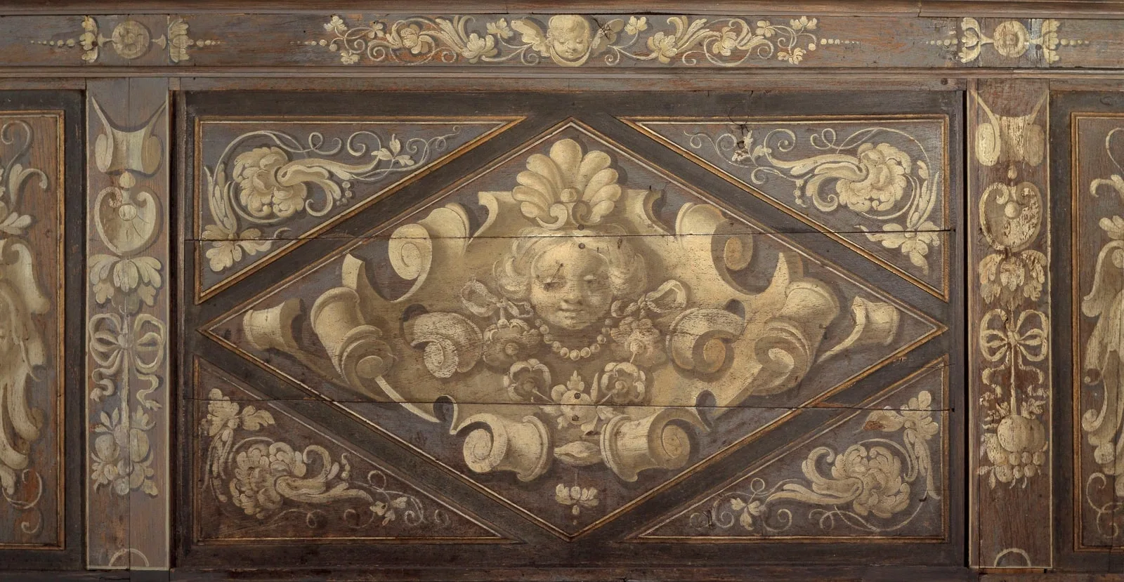 Panneau de bas-lambris à compartiment en losange inscrit dans un rectangle. Dans le losange, tête féminine, fleurs, perles et rubans se détachant sur un motif de cuirs.